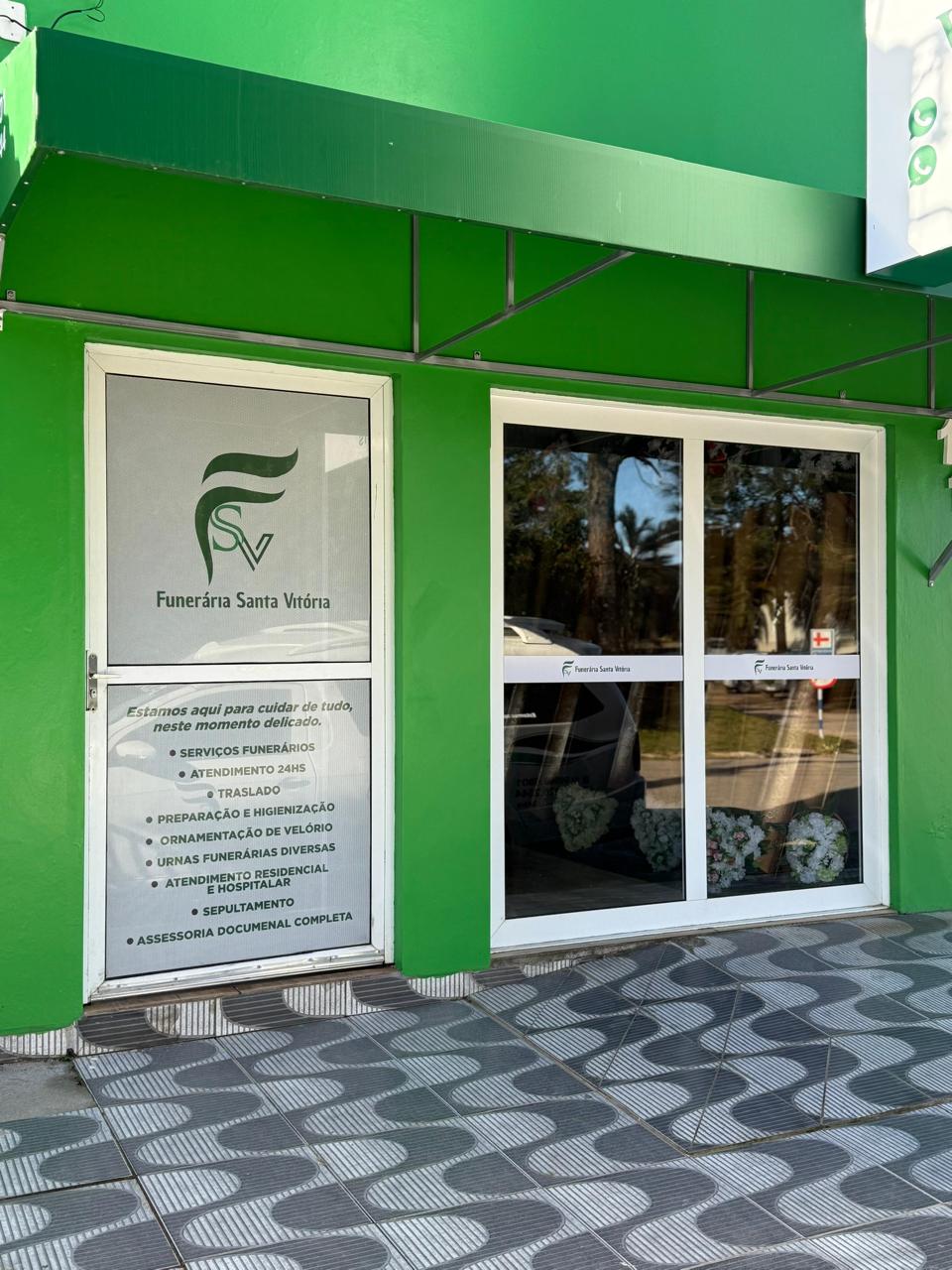 fachada de funerária com paredes e toldo verdes, porta de vidro com logo e lista de serviços e coroas de flore